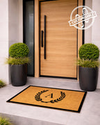 Doormat with a monogram in front of a modern wooden door with plants on either side. [VARIANT: SINGLE INITIAL / BORDER]