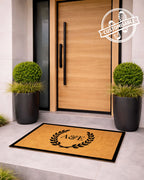 Doormat with a monogram in front of a modern wooden door with plants on either side. [VARIANT: TWO INITIALS / BORDER]