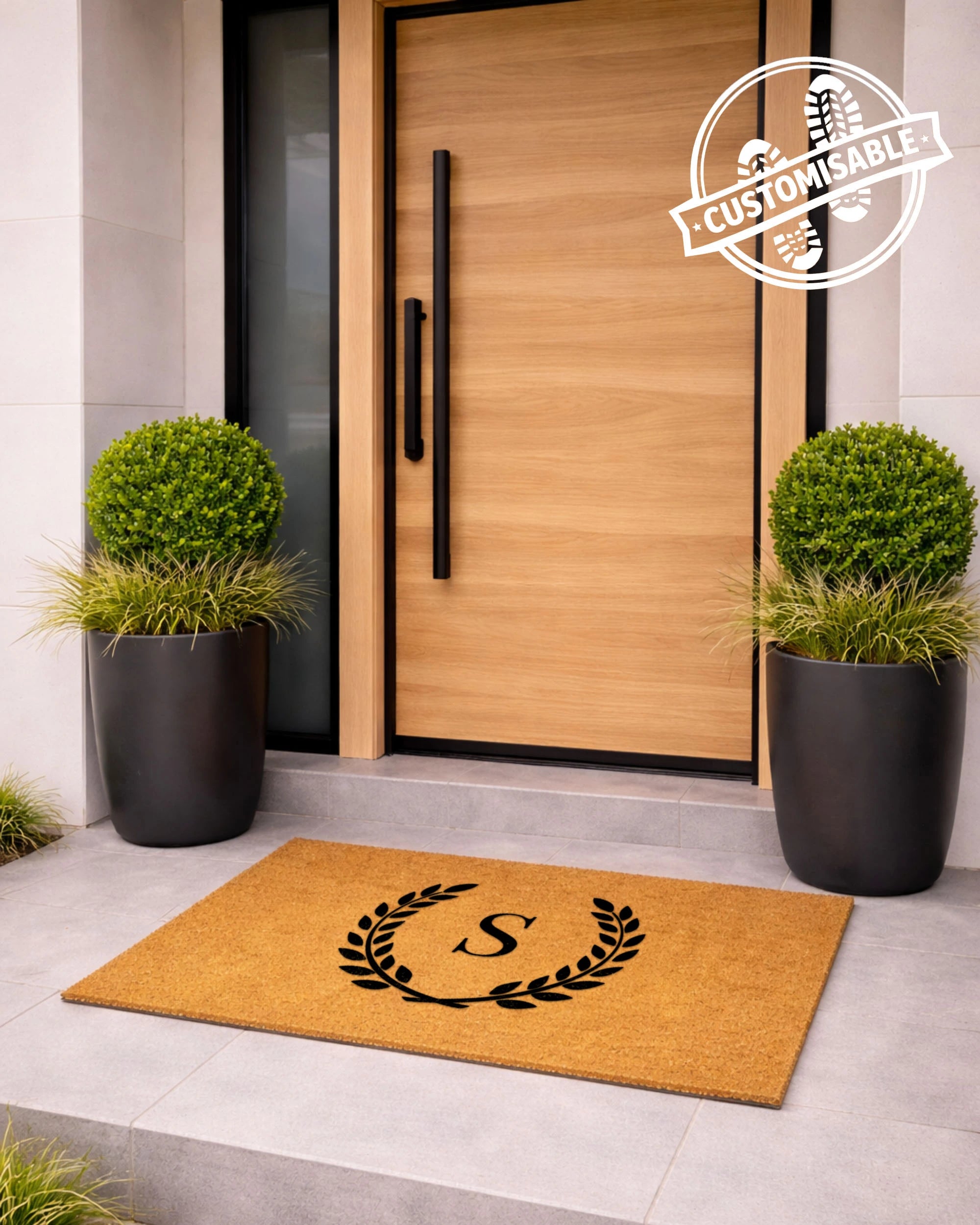 Doormat with a monogram in front of a modern wooden door with black handles, flanked by two potted plants. [VARIANT: SINGLE INITIAL / NO BORDER]