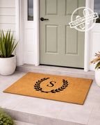 Doormat with monogram design in front of a door, featuring a customizable label. [VARIANT: SINGLE INITIAL / NO BORDER]