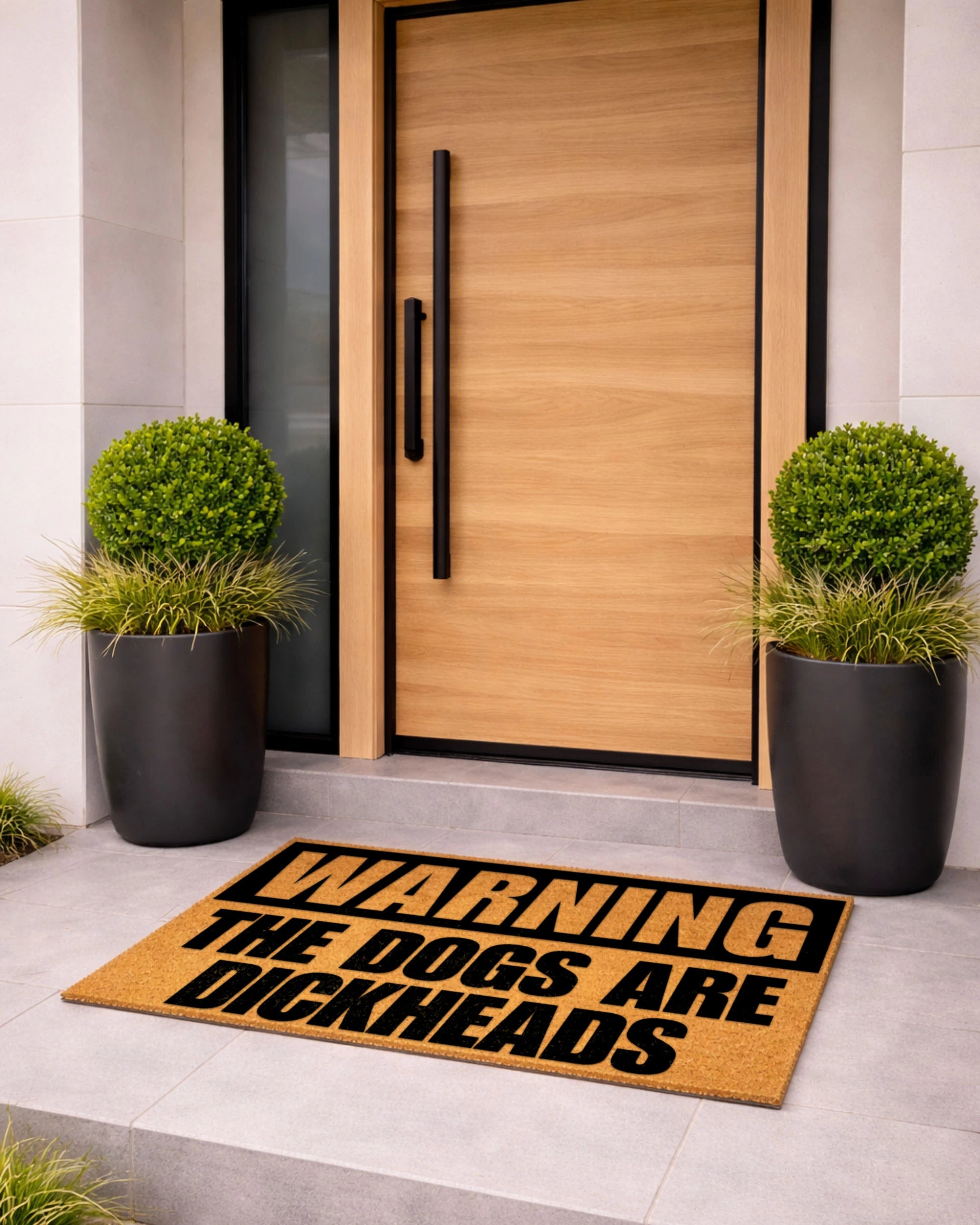 Doormat with humorous text in front of a modern wooden door with black handles, flanked by two potted plants. [VARIANT: DOGS / DICKHEADS]