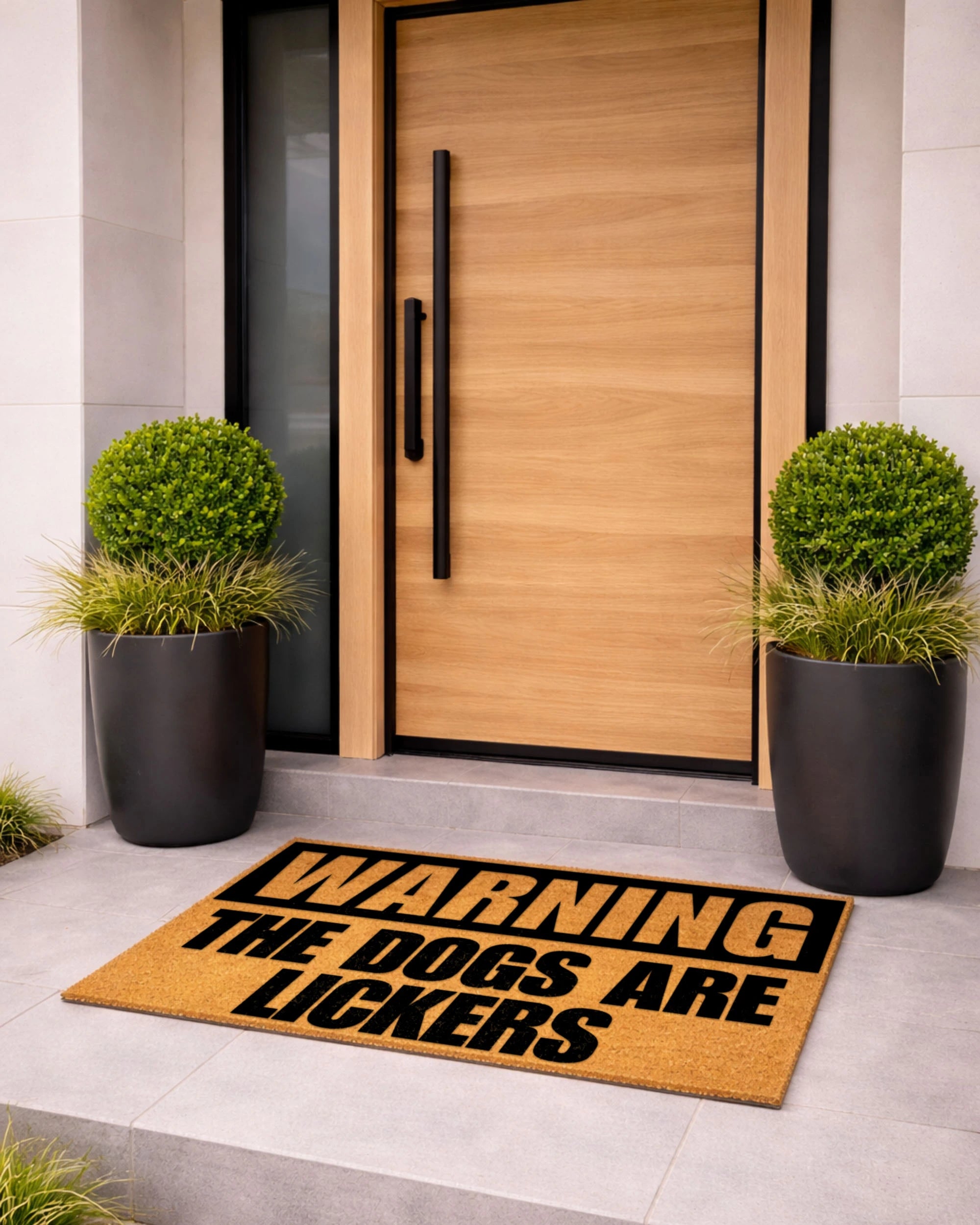 Doormat with humorous text in front of a modern wooden door with black handles, flanked by two potted plants. [VARIANT: DOGS / LICKERS]