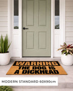 Doormat with humorous text in front of a green door on a house exterior. [VARIANT: DOG / DICKHEAD]