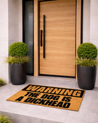 Doormat with humorous text in front of a modern wooden door with black handles. [VARIANT: DOG / DICKHEAD]