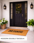 Front door with a doormat featuring 'NO CUNTS' text, flanked by potted plants. [variant: block]