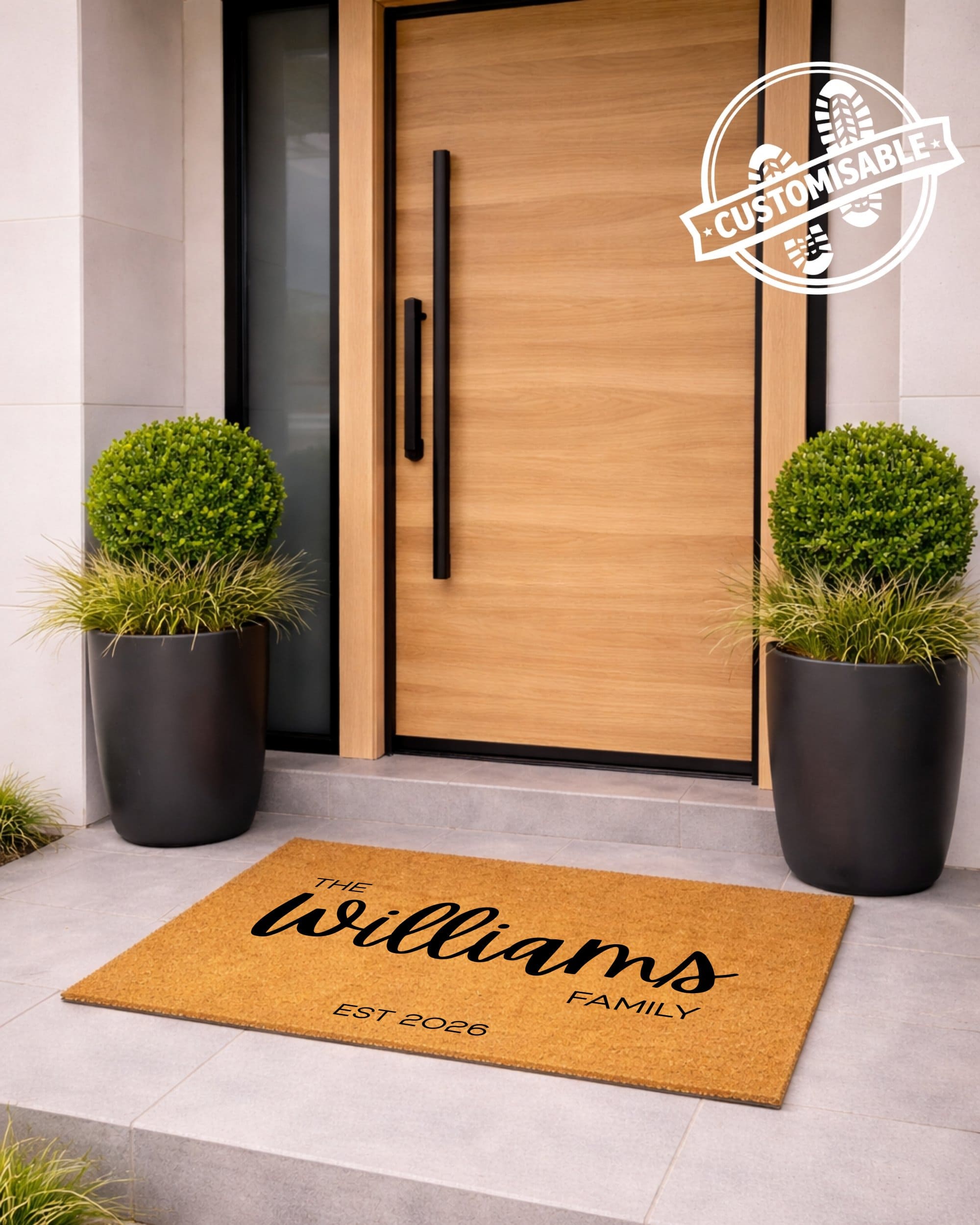 Doormat with 'The Williams Family' text in front of a modern wooden door with plants on either side. [VARIANT: STANDARD-EST]