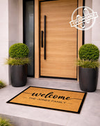 Doormat with 'welcome THE JONES FAMILY' text in front of a modern wooden door with plants on either side. [variant: BORDER]