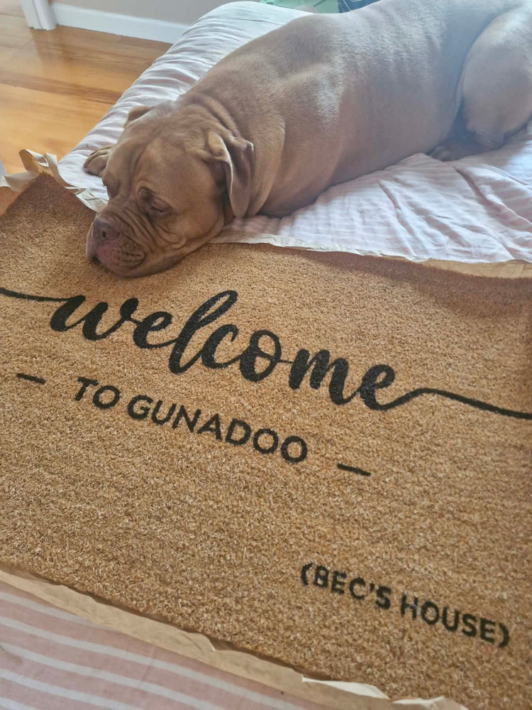 Dog lying on a doormat with 'welcome to gunadoo Bec's house' text