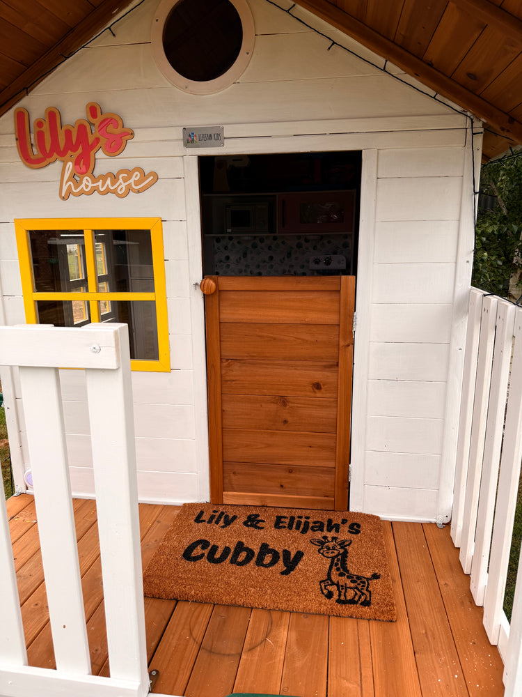 Wooden playhouse with a door and a doormat featuring text and graphics.