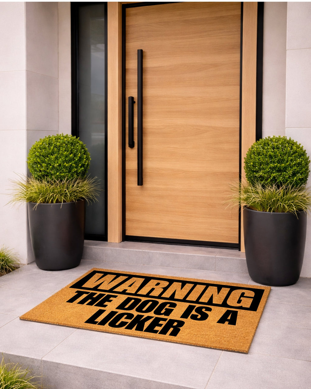 Doormat with humorous text in front of a modern wooden door with black handles, flanked by two potted plants. [variant: dog-licker]