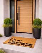 Doormat with Please take off your shoes my kids lick the floor in front of a modern wooden door with black handles, flanked by two potted plants.