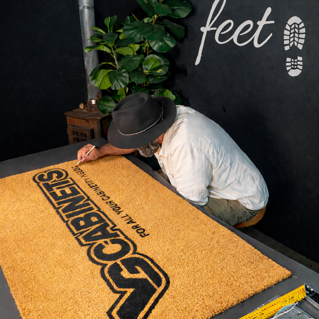 Person adjusting a doormat with a logo on a table, against a black wall with 'Mudd Feet' text.