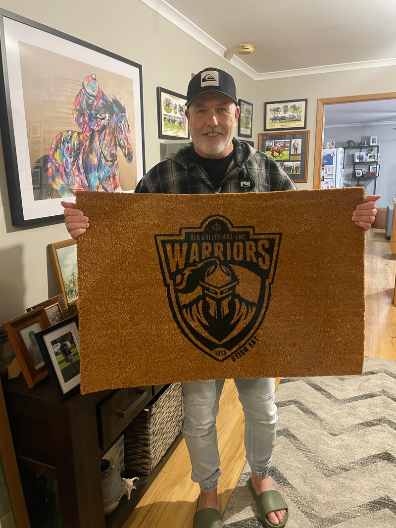 Person holding a doormat with a logo in a room with framed pictures on the wall.