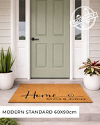 Doormat with 'Home Emma & Joshua' text in front of a green door on a house exterior.