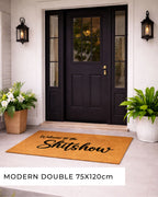 Doormat with 'NO CUNTS' text in front of a black door with glass panels, flanked by potted plants.