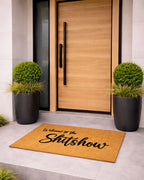 Doormat with 'Welcome to the Shitshow' text in front of a wooden door with black handles.